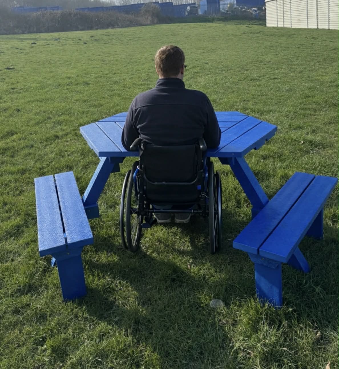 Person in a wheelchair sits at a bright blue outdoor picnic table in a grassy park, back facing the camera (outdoor setting).