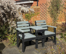 Outdoor seating area with gray wooden benches placed on a black platform, set against a brick wall with a lattice trellis and white flowering shrub to the left.